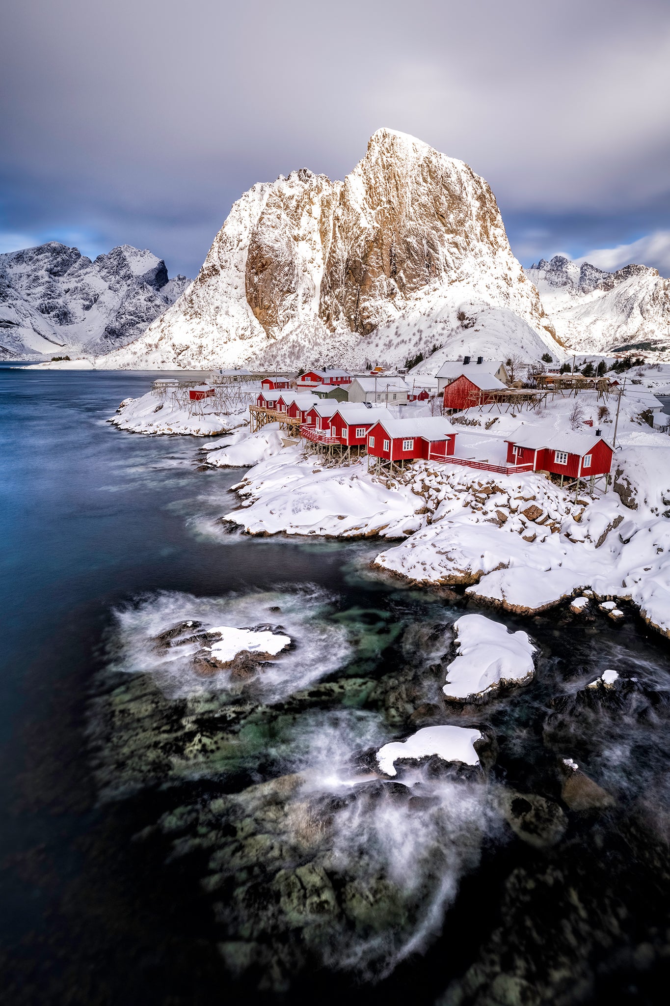 Red cabins nestled among snow-covered mountains with a body of water in the foreground. Lofoten. Hamnoy