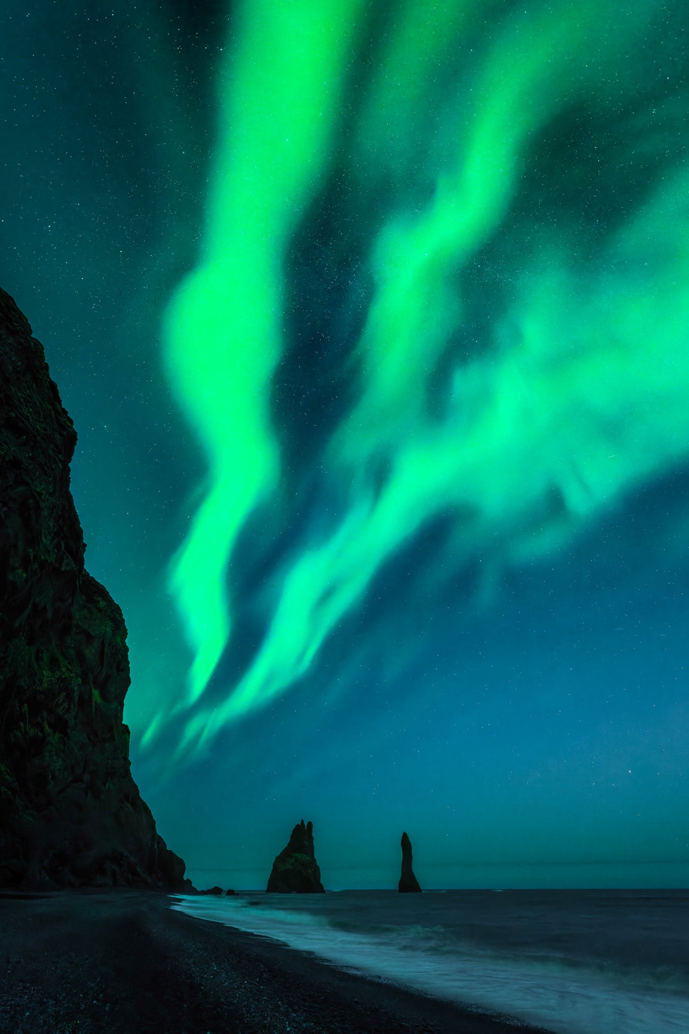 Reynisdrangar. Aurora borealis over a rocky landscape with a path leading to a body of water.
