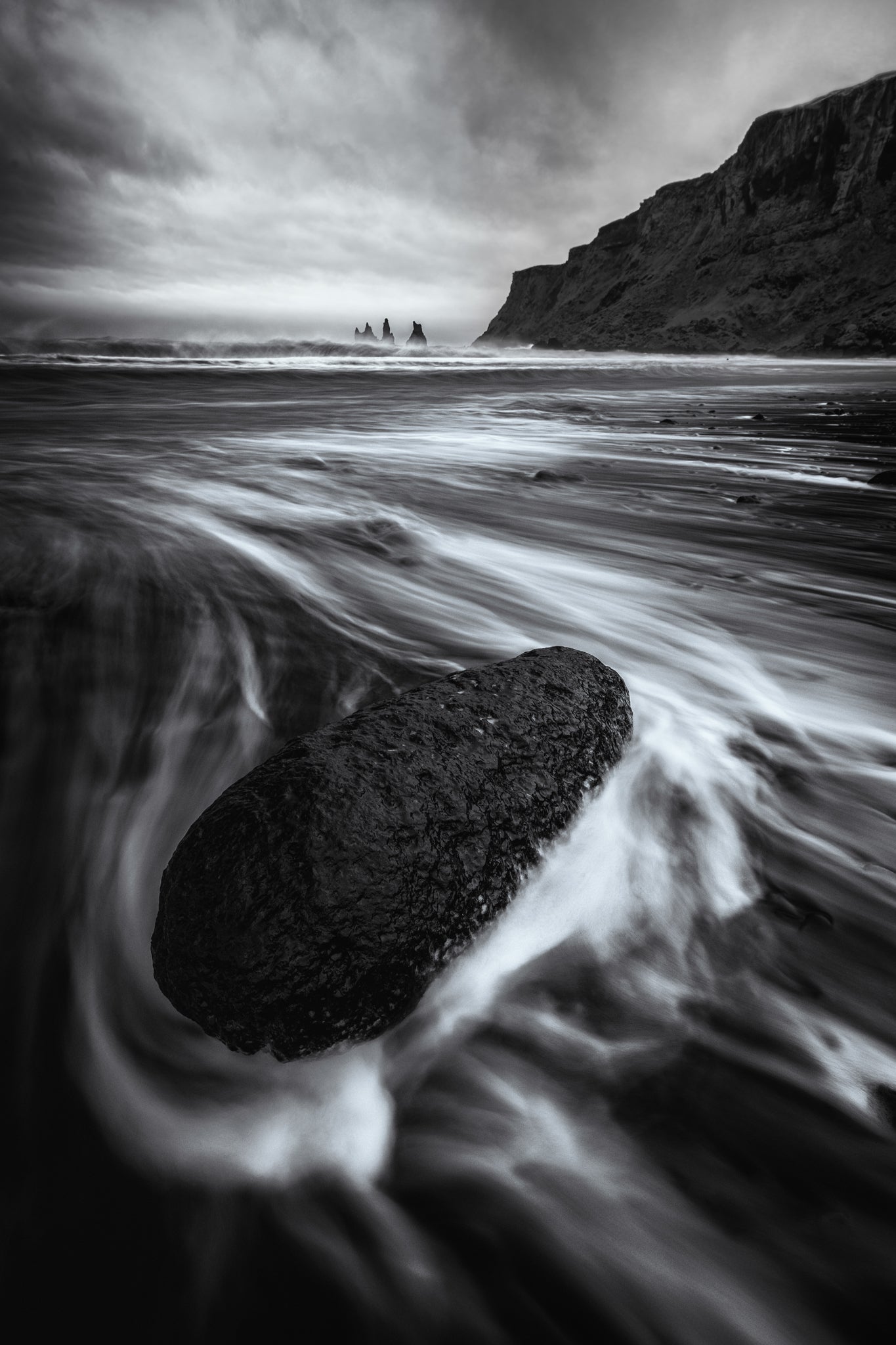 Vik. Black and white photograph of a rock in water with a mountainous landscape in the background