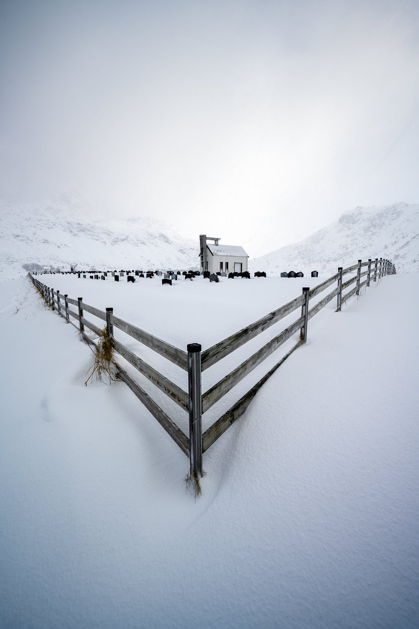 Snow-covered landscape with a wooden fence and a small building in the distance.