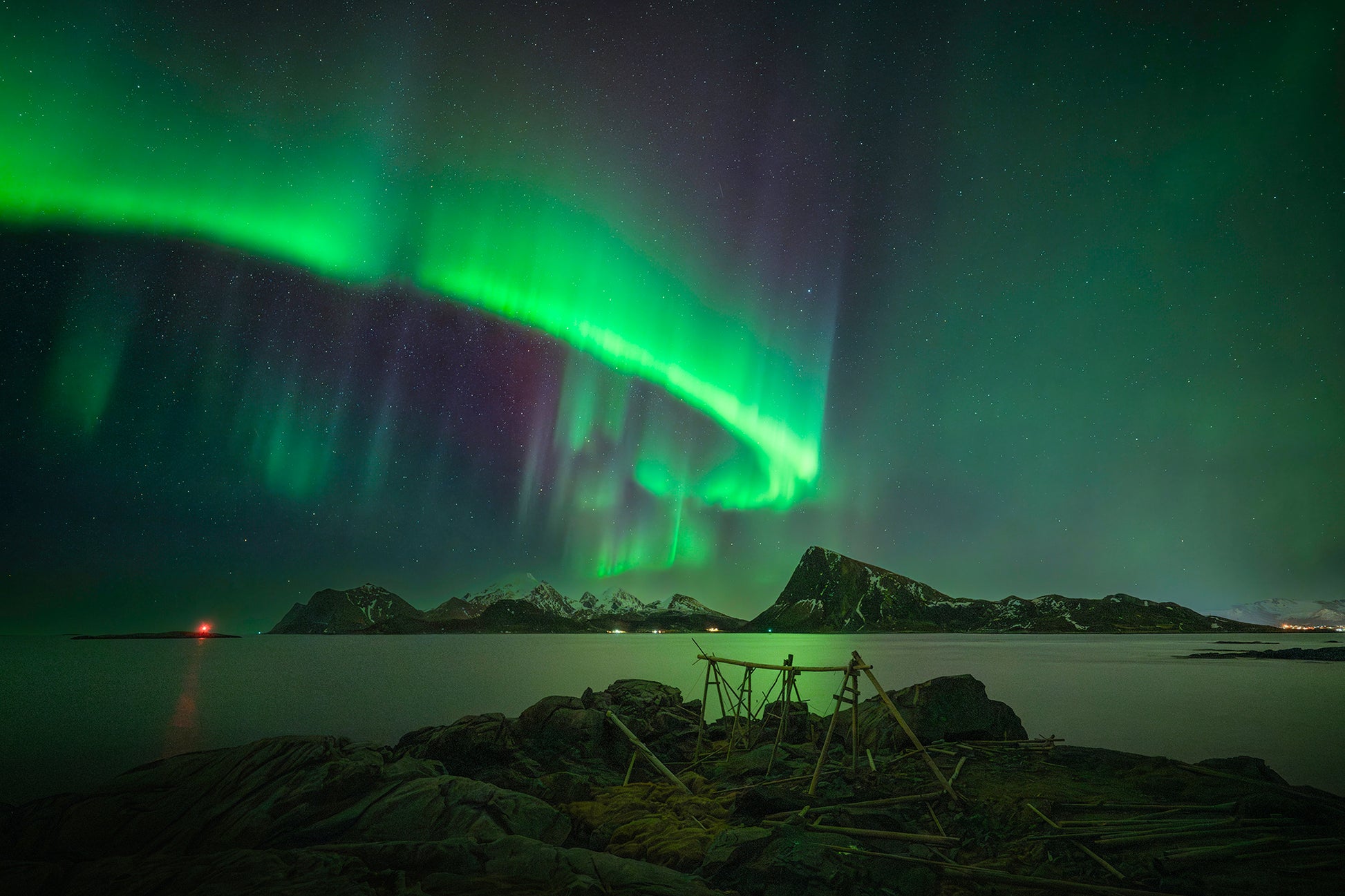 Aurora borealis over a body of water with rocks and a small structure in the foreground.