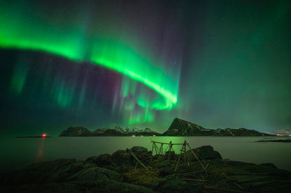 Aurora borealis over a body of water with rocks and a small structure in the foreground.
