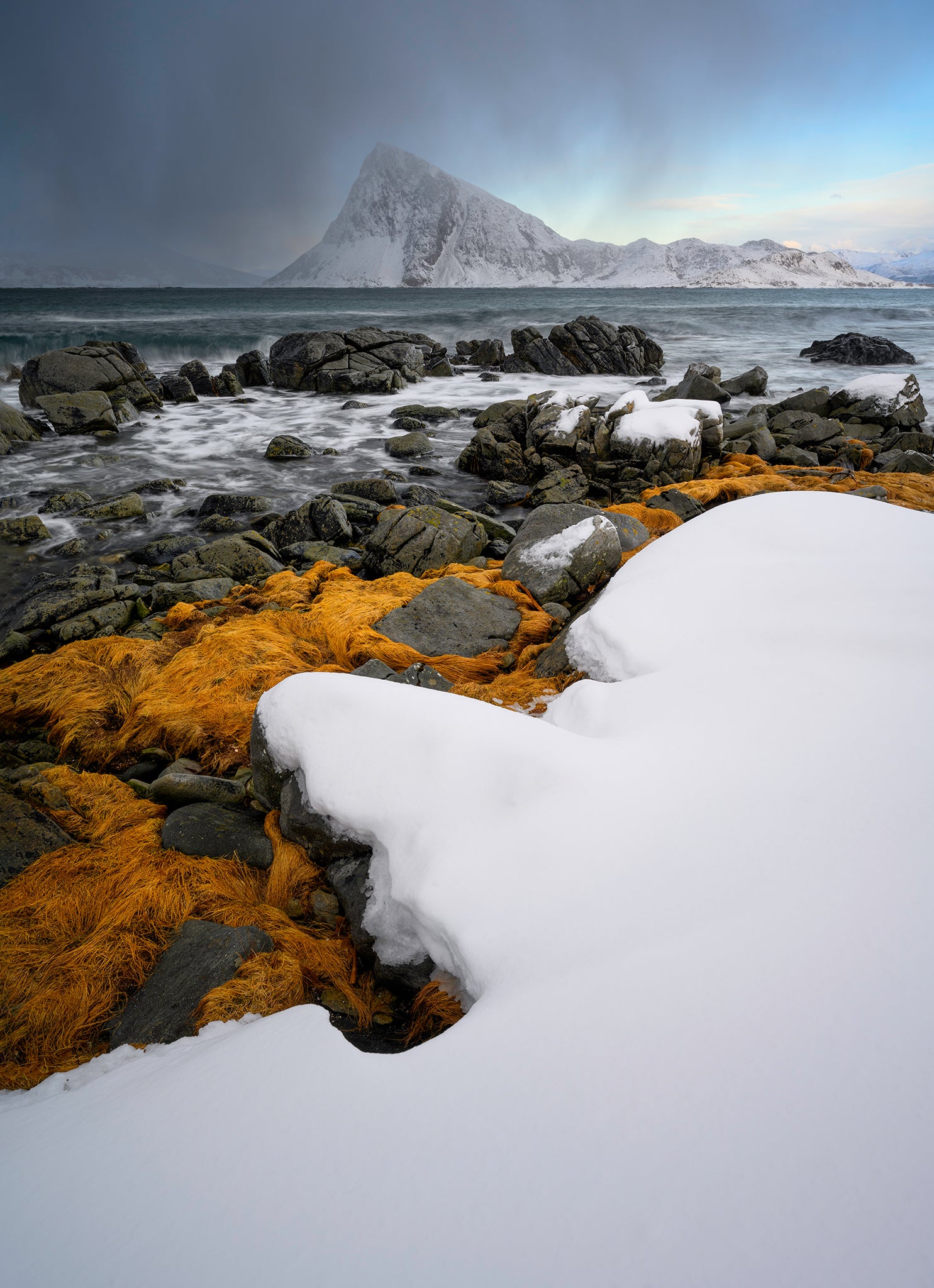 Snow-covered rocks with orange lichen on a coastal landscape with a mountain in the background.