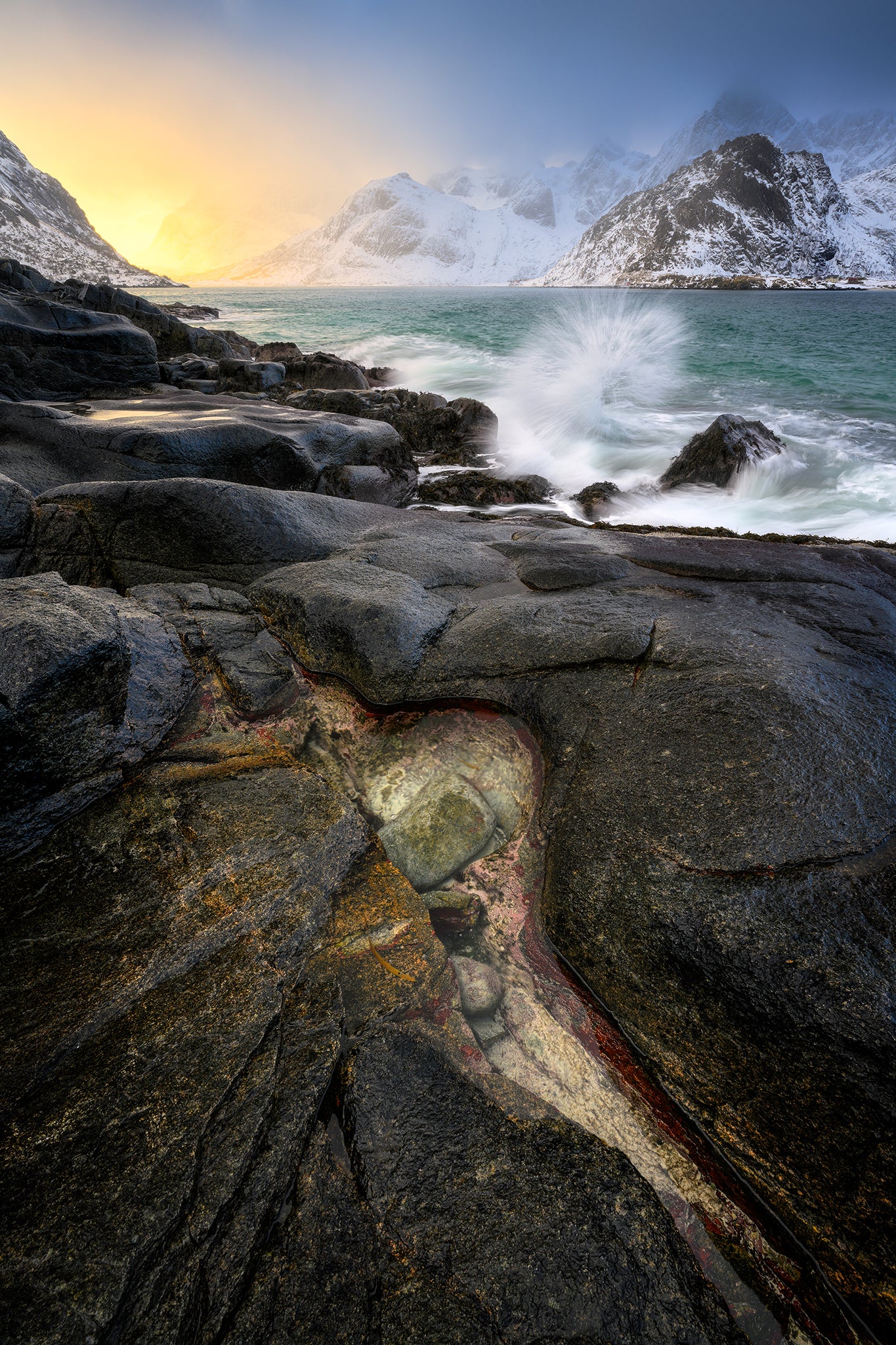 Rugged coastal landscape with waves crashing against rocks and snow-capped mountains in the background.