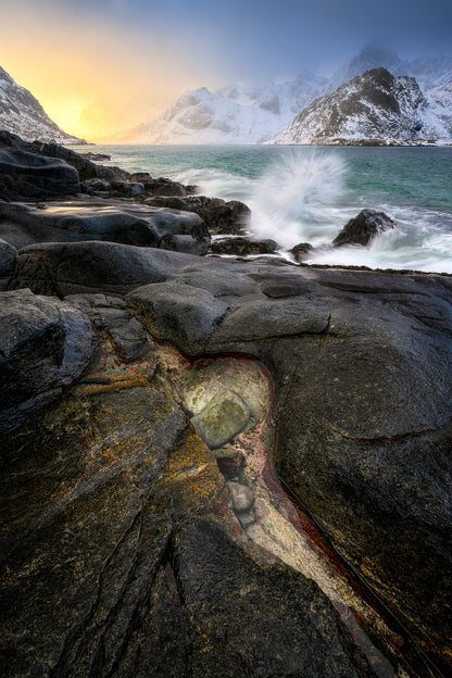 Rugged coastal landscape with waves crashing against rocks and snow-capped mountains in the background.